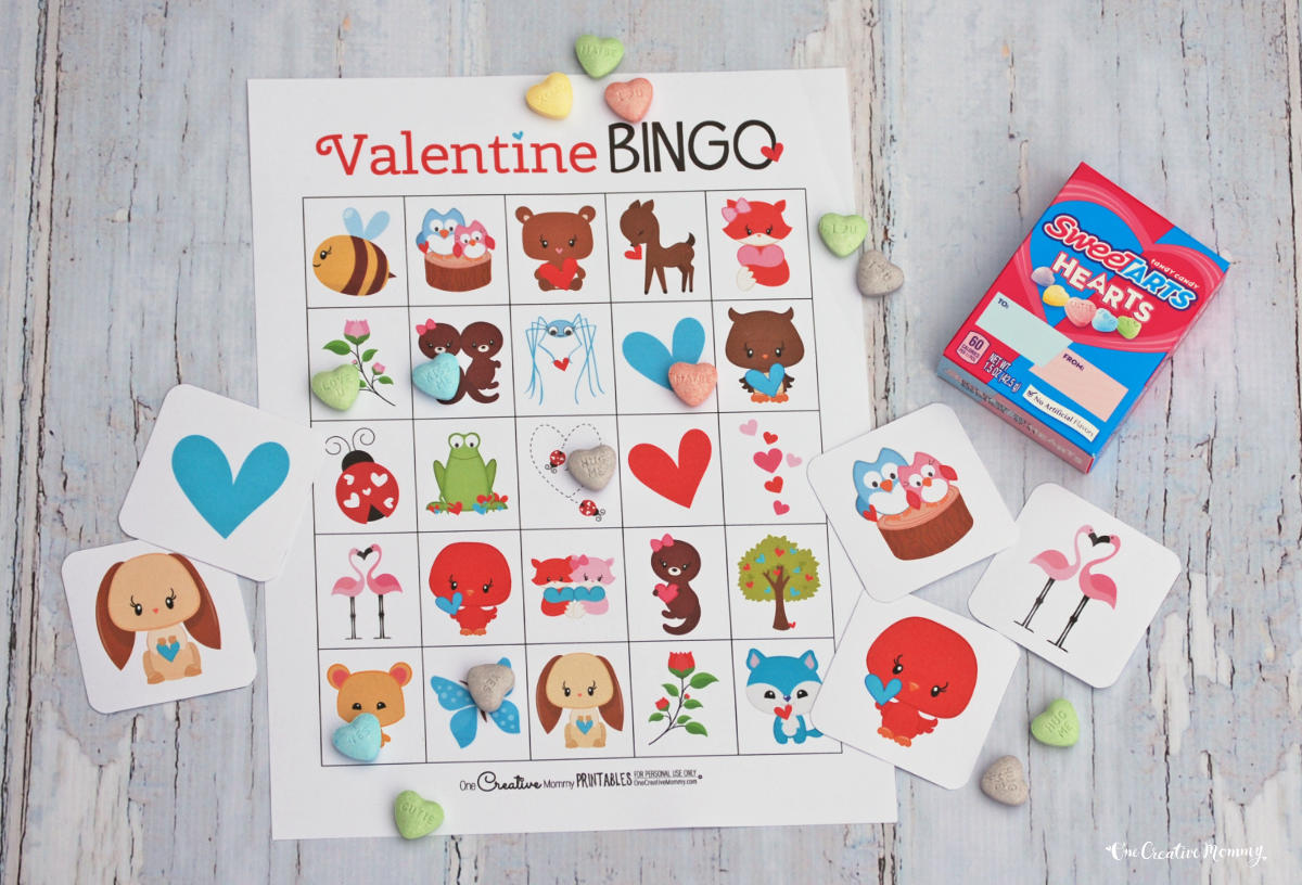 A Valentine's Day bingo card is shown laying on a white wooden tabletop. Five calling cards from the game are laid out across the page. Sweethearts candies are scattered across the game boards. Elements on the game include cute cartoon animals, hearts, flowers, etc.