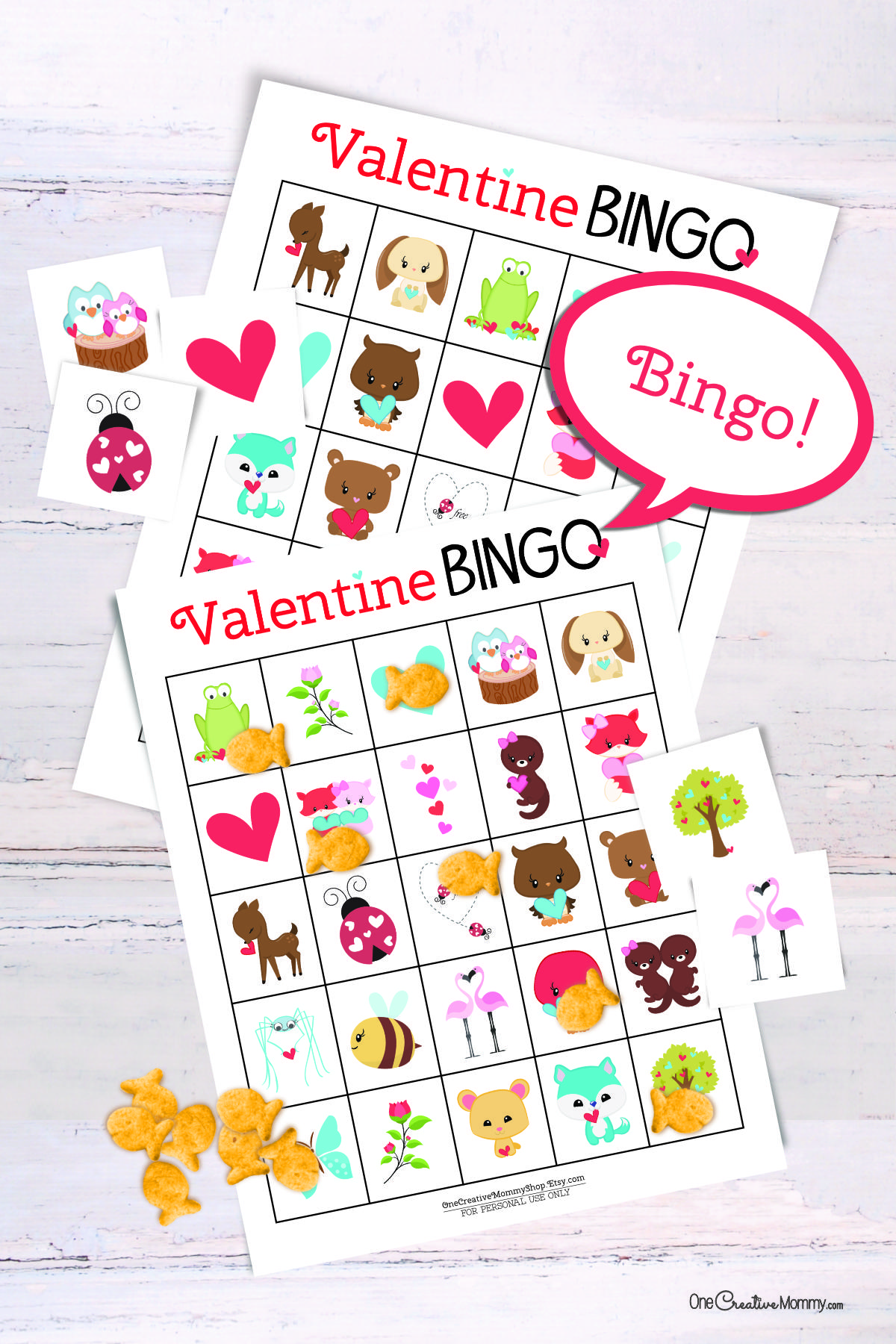 2 Valentines Day bingo cards are laid on top of each other on a white table. There is a small pile of goldfish crackers. Six calling cards are scattered across the game, and several spots are marked with crackers. A word bubble says Bingo!