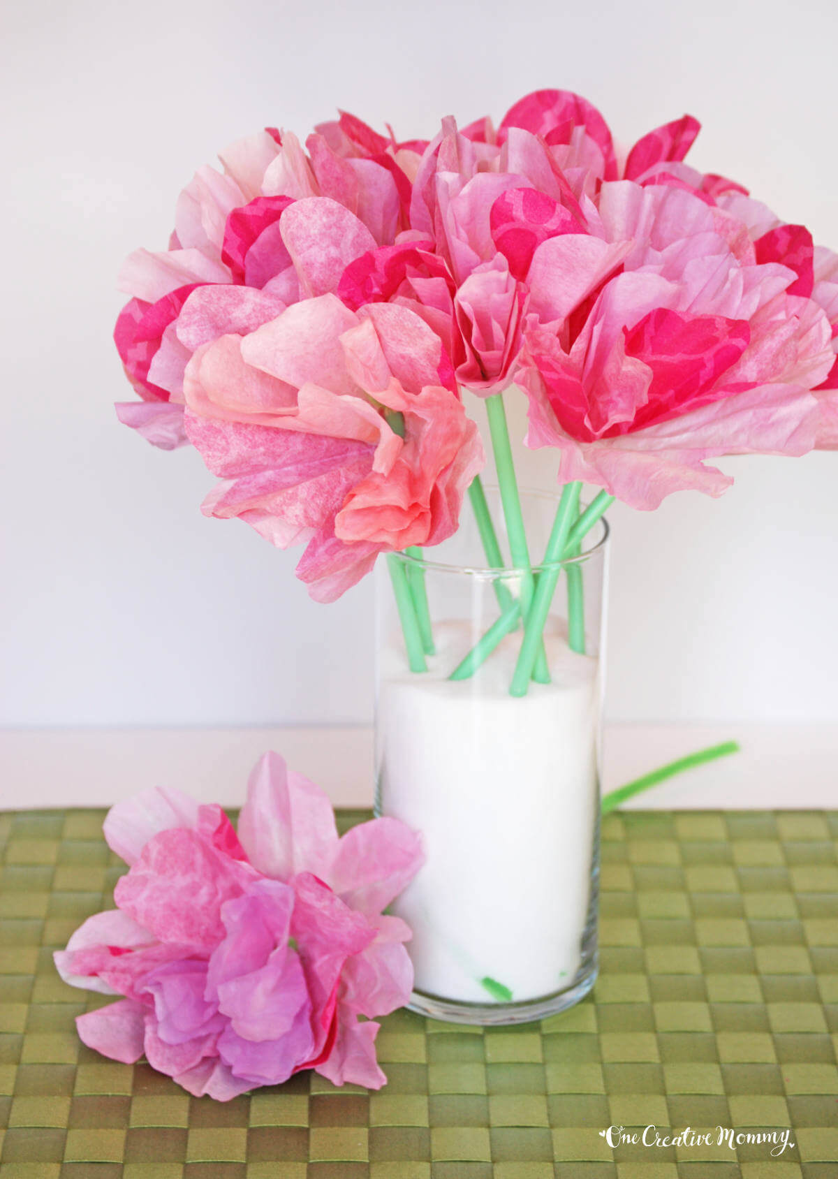 A clear vase filled with white salt holds several pink coffee filter flowers with green stems made from drinking straws.
