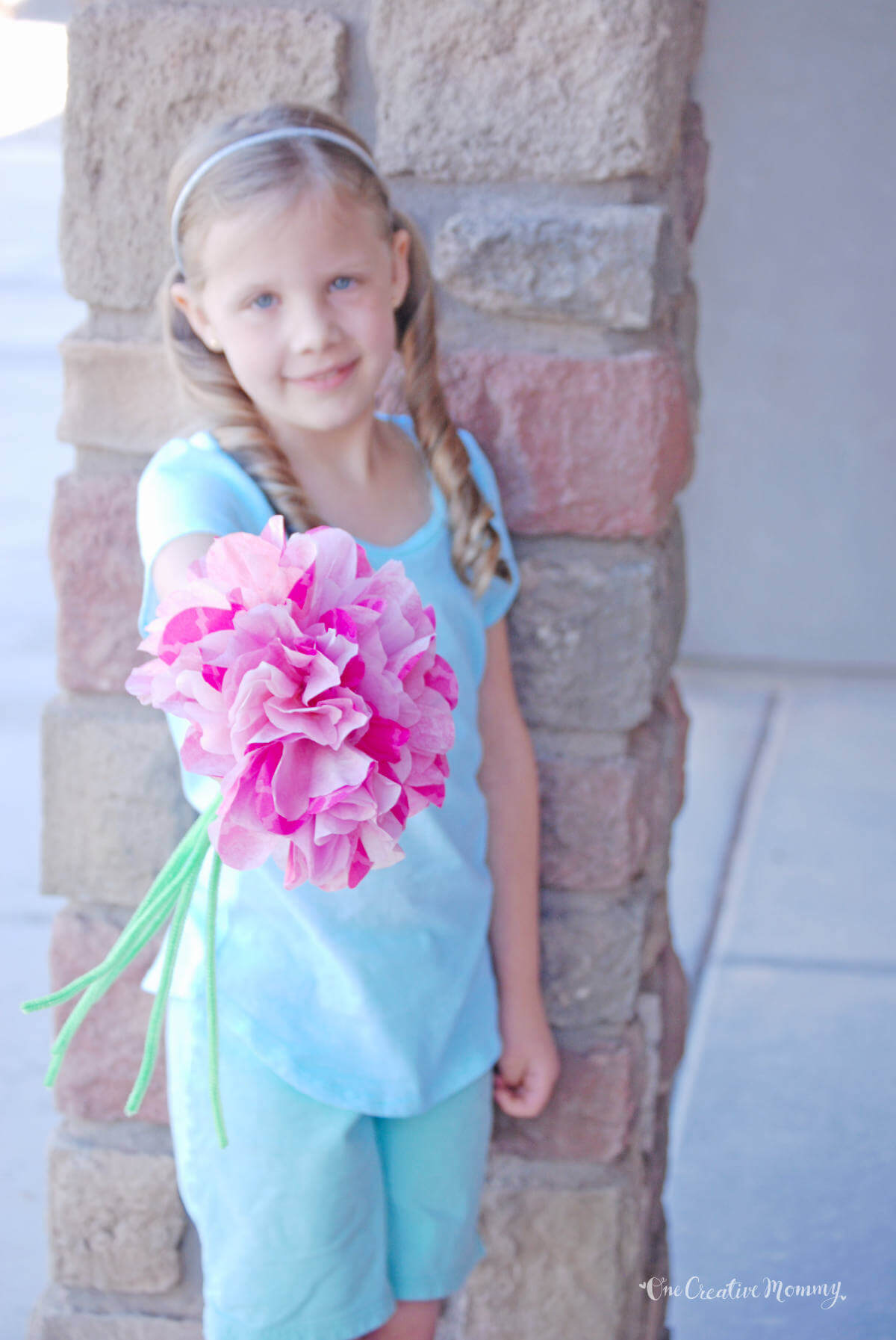 A little girl wearing light blue shorts and t-shirt stands in front of a brick pillar. She is smiling and holding a bouquet of pink coffee filter flowers with green chenille stems out toward the camera.
