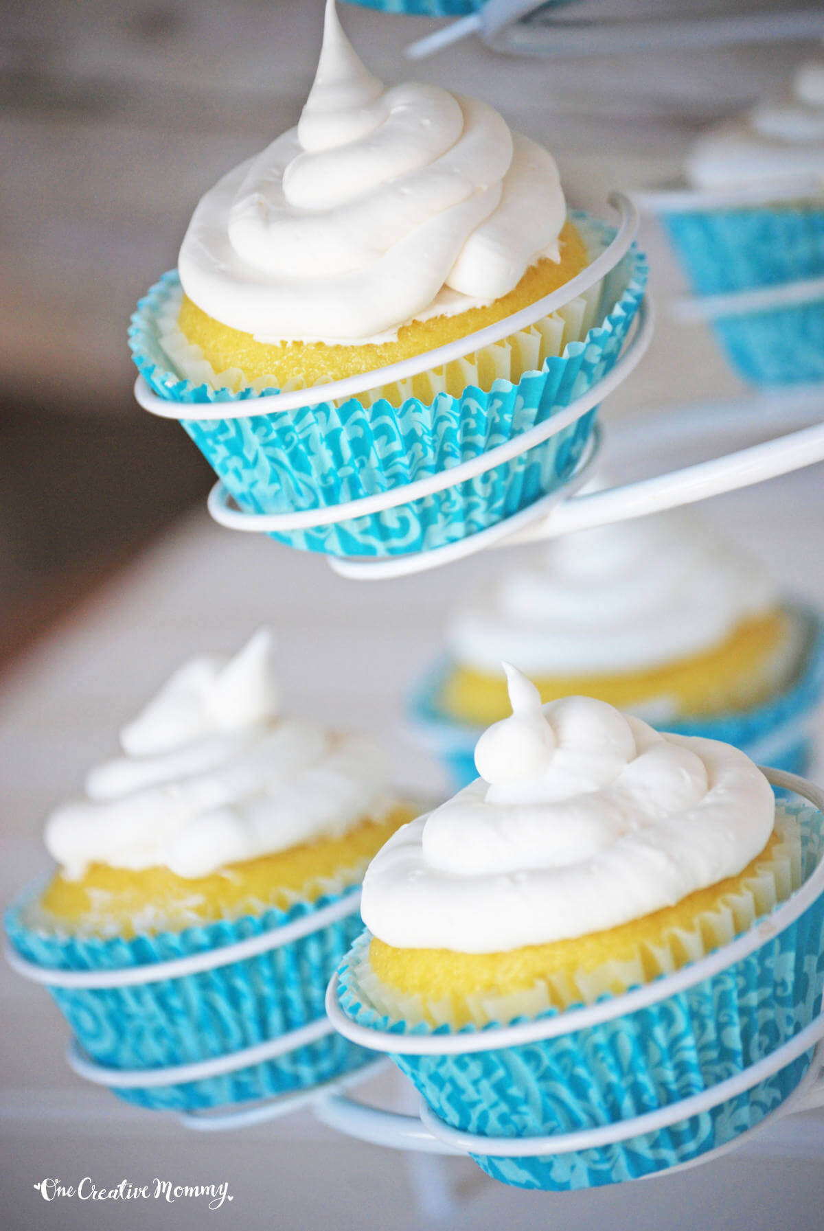Several delicious gluten free lemon pound cake cupcakes arranged on a spiral cupcake stand. They are bright yellow, frosted with white frosting, and nestled inside blue cupcake liners.