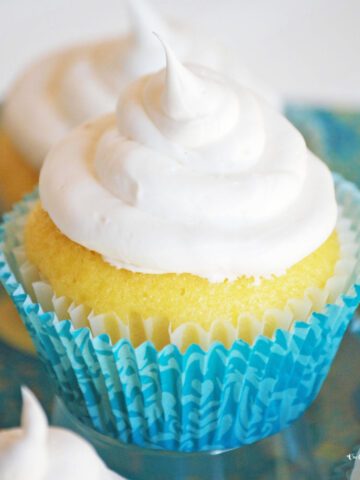 Several delicious gluten free lemon pound cake cupcakes arranged on a pretty plate. They are bright yellow, frosted with white frosting, and nestled inside blue cupcake liners.
