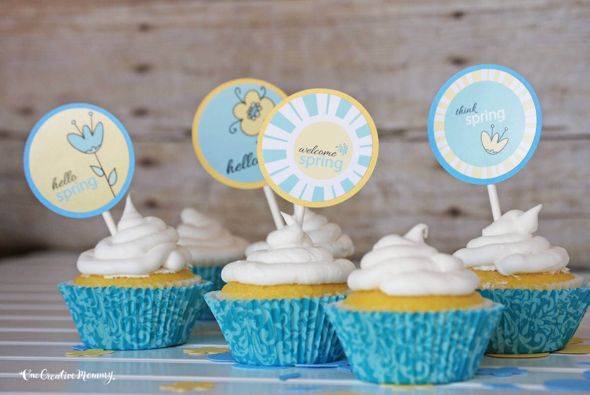 Cute spring cupcake toppers spear several delicious gluten free lemon pound cake cupcakes arranged across a table. The toppers are pale yellow and blue and say, "Welcome Spring," "Think Spring," "Hello Spring," and "Hello." The cupcakes are bright yellow, frosted with white frosting, and nestled inside blue cupcake liners.