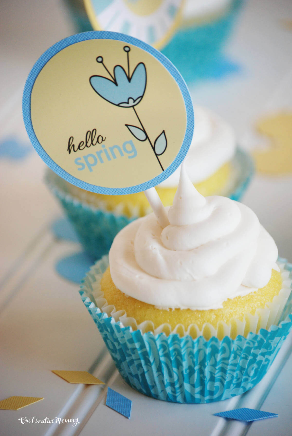 A cute spring cupcake topper spears a delicious-looking gluten free lemon pound cake cupcake. The topper is pale yellow and blue and says, "Hello Spring" The cupcake is bright yellow, frosted with white frosting, and nestled inside a blue cupcake liner.