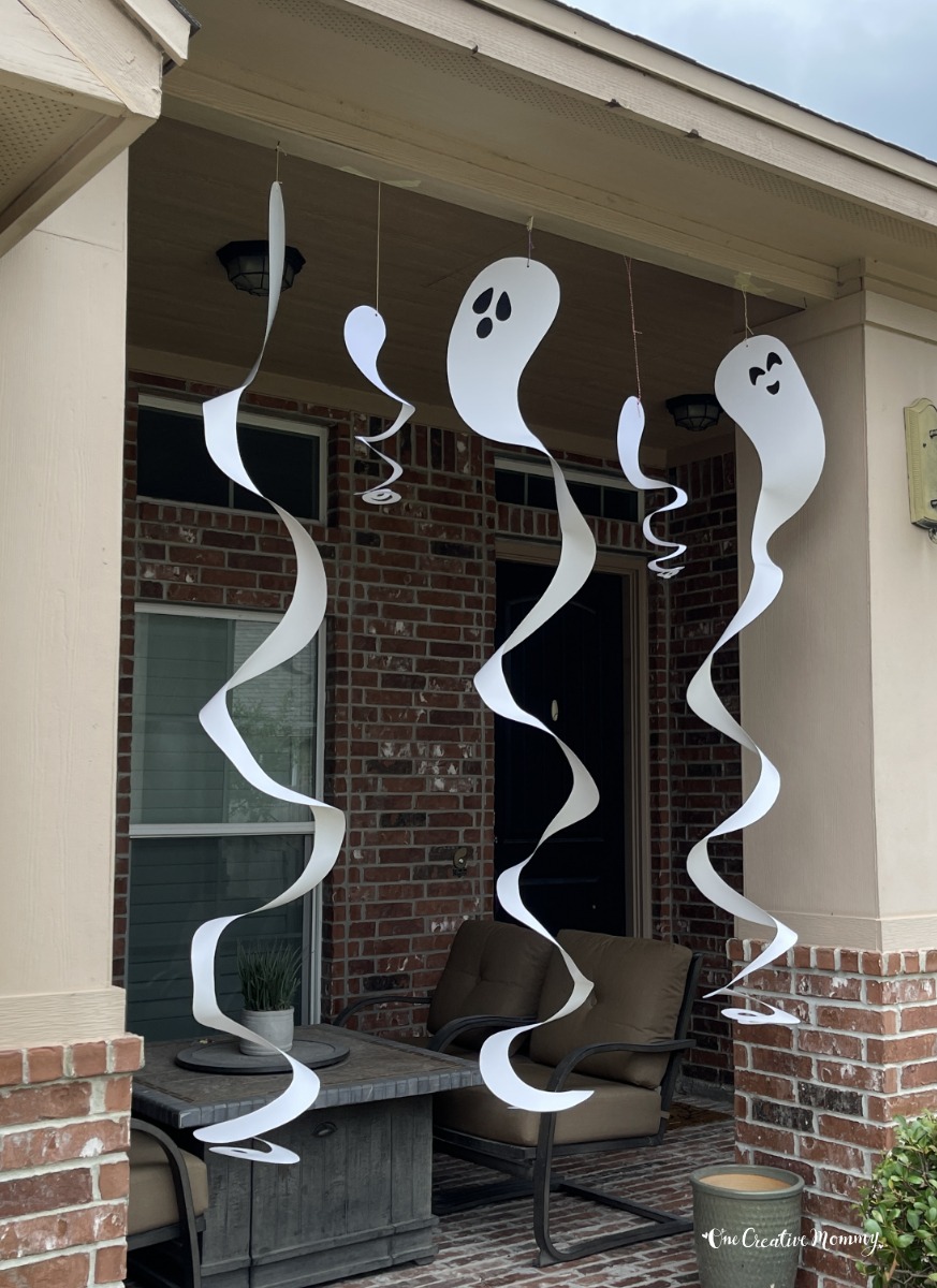 Spiral ghosts of several sizes and made from white cardboard hang from the roof of a home's front porch. The ghosts have creepy faces, and they are spinning in the wind.