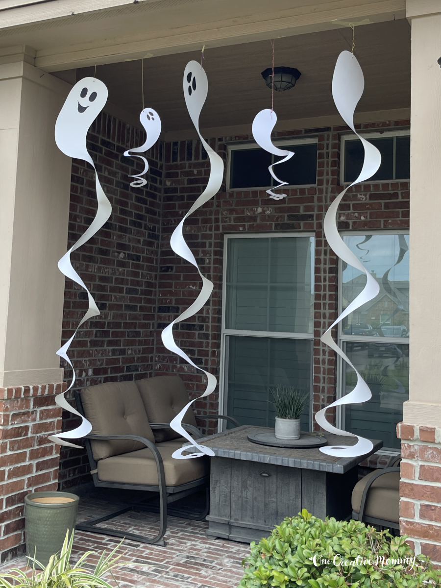 Spiral ghosts of several sizes and made from white cardboard hang from the roof of a home's front porch. The ghosts have creepy faces, and they are spinning in the wind.