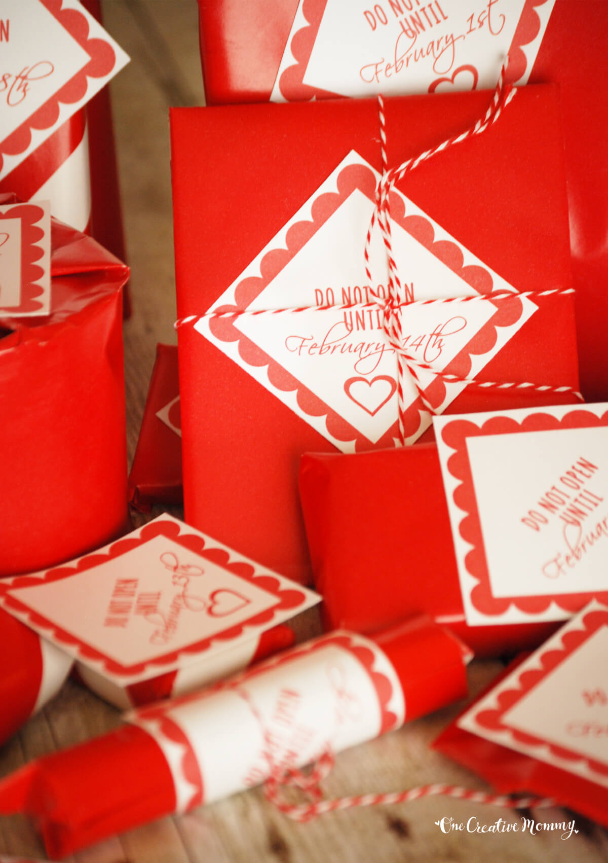 14 Valentine's Day gifts wrapped in red paper with a Do Not Open Until cards attached to each gift with red baker's twine. The gifts are piled up on top of a wooden table.