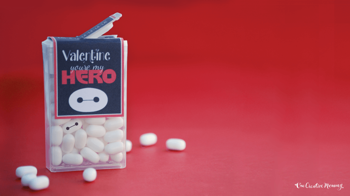 An open package of white Tic Tac candies is shown on a red background. Several tic tacs have spilled out onto the table. The logo is covered by a tag that says, Valentine, you're my hero, with a picture of Baymax.