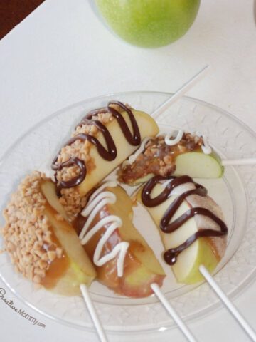 5 slices of red and green apples speared on lollipop sticks and covered with caramel, nuts, toffee, and drizzled chocolate sit on a clear plate