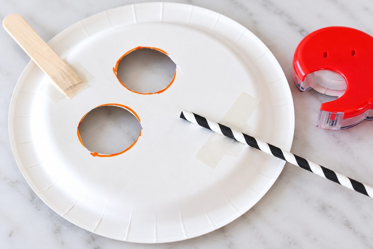 Back of a paper plate pumpkin mask showing a wooden stem and black-and-white striped straw handle secured with tape.