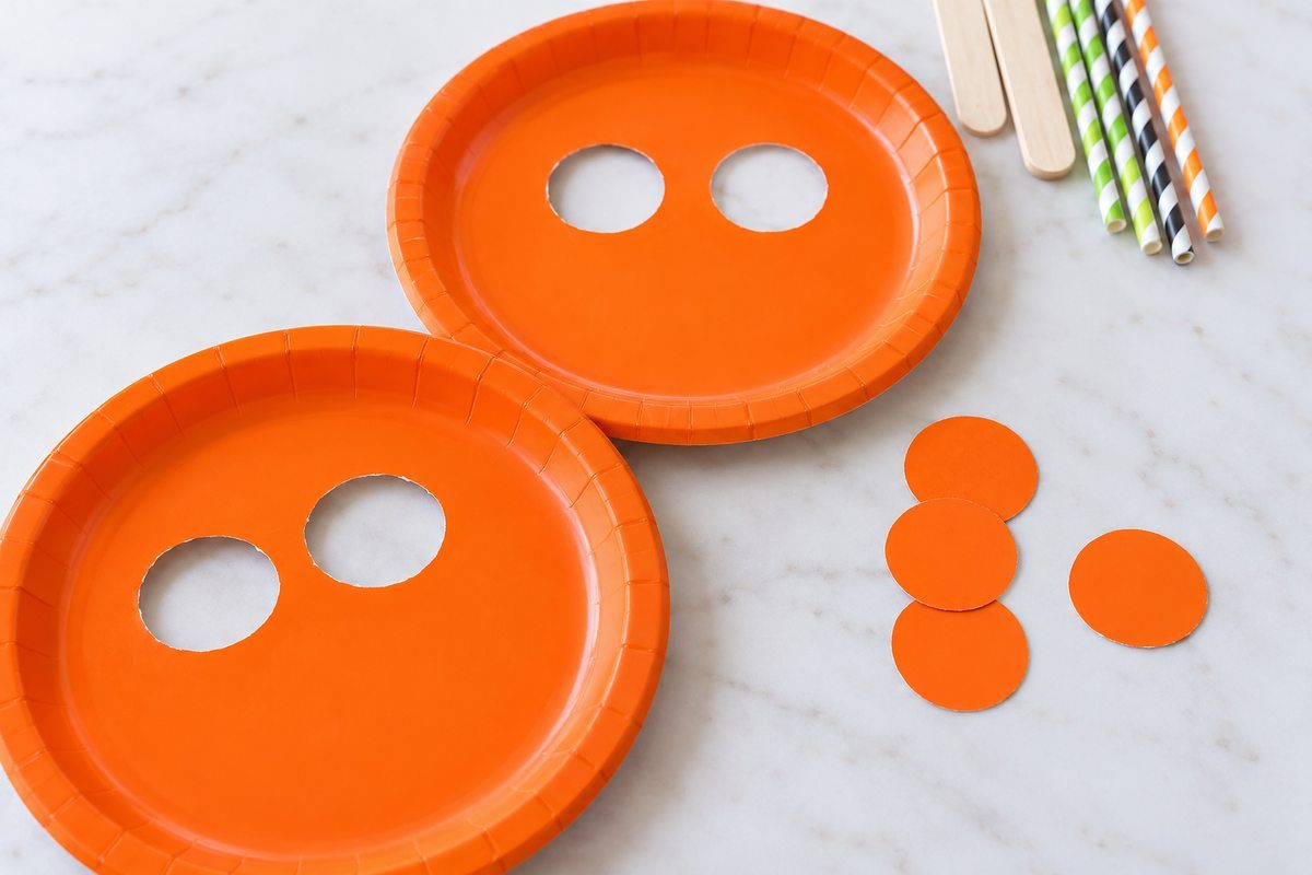 Two orange paper plates with circular eye holes cut out, surrounded by paper cutouts, craft sticks, and striped straws.