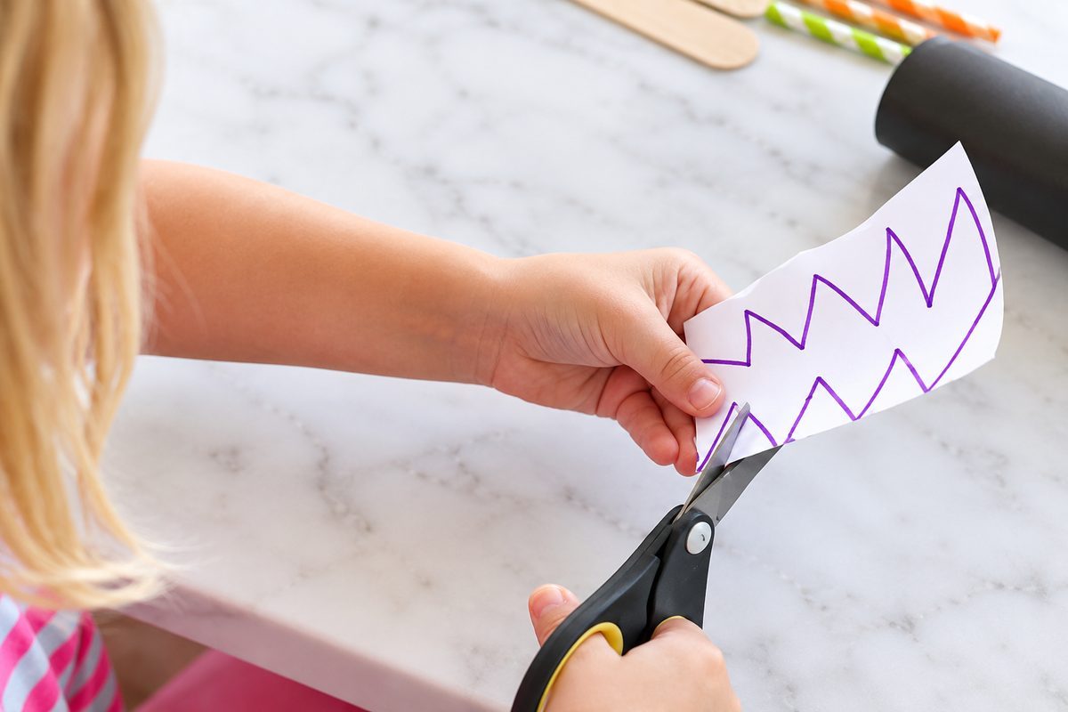 Child using scissors to cut out a zigzag jack-o'-lantern mouth shape for a paper plate pumpkin mask.