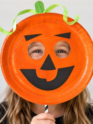 Girl peeking through the eye holes of a smiling orange paper plate pumpkin mask decorated with a green ribbon stem.