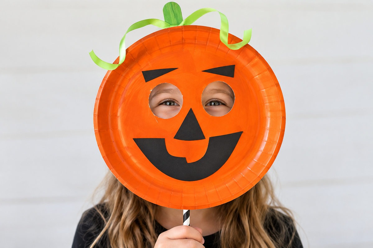 Girl peeking through the eye holes of a smiling orange paper plate pumpkin mask decorated with a green ribbon stem.